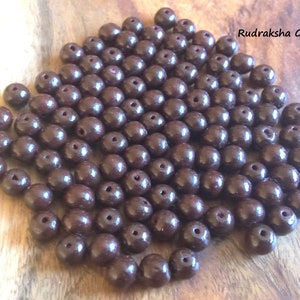 May include: A pile of dark brown round beads, likely made of wood, with a small hole in the center of each bead. Rudraksha Gems is written in white text on a brown background in the upper right corner of the image.