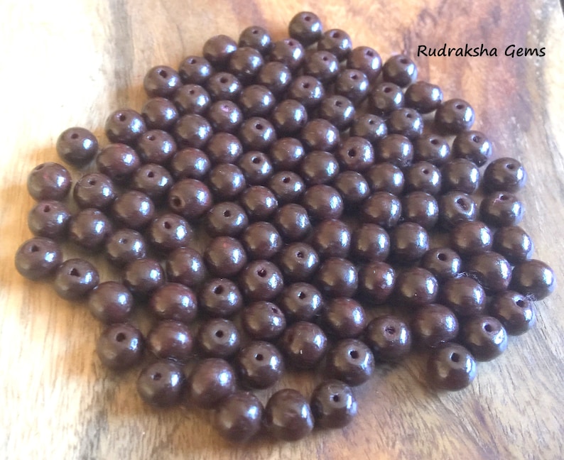 May include: A pile of dark brown round beads, likely made of wood, with a small hole in the center of each bead. Rudraksha Gems is written in white text on a brown background in the upper right corner of the image.