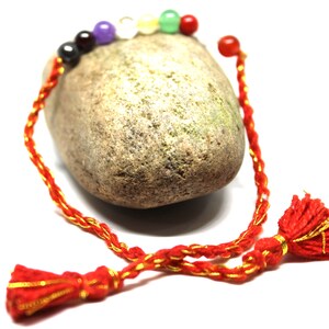 May include: Red string bracelet with seven colorful beads and gold accents, tied with red tassels.
