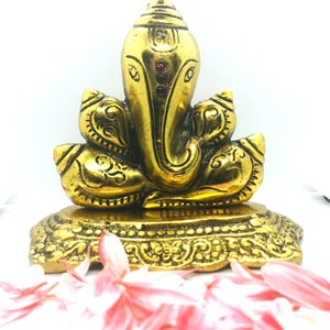 Puede incluir: Una estatua dorada de la deidad hind&uacute; Ganesha, con detalles intrincados y acentos rojos. La estatua est&aacute; sobre una base decorativa, con p&eacute;talos de flores rosas en primer plano. El texto "Rudraksha Gems" es visible en la parte inferior.