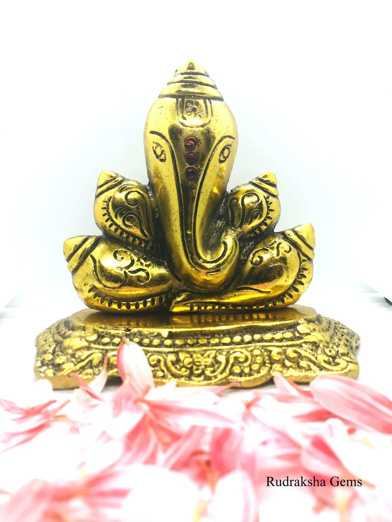 Puede incluir: Una estatua dorada de la deidad hind&uacute; Ganesha, con detalles intrincados y acentos rojos. La estatua est&aacute; sobre una base decorativa, con p&eacute;talos de flores rosas en primer plano. El texto "Rudraksha Gems" es visible en la parte inferior.