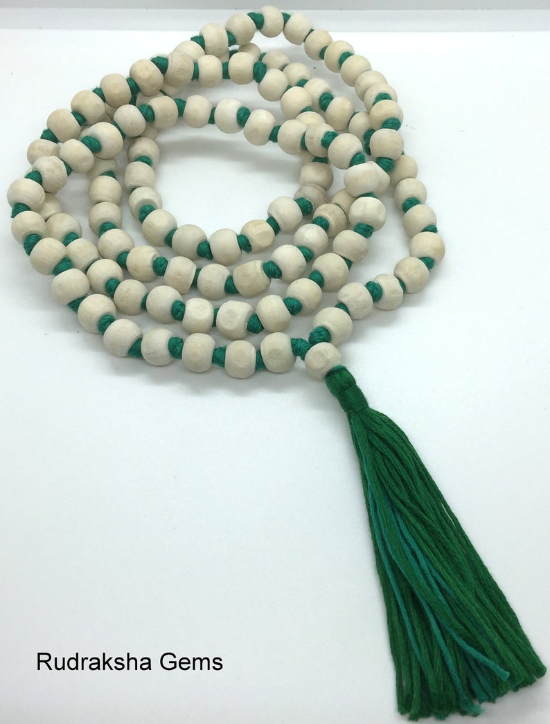 May include: A string of light-coloured, round wooden beads knotted onto a vibrant green cord, arranged in a circular pattern. The necklace features a long, matching green tassel at the end. The text Rudraksha Gems is printed in the bottom corner.