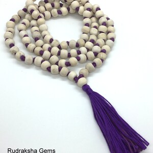 May include: A long necklace featuring natural light-coloured wooden beads strung on a vibrant purple cord, arranged in multiple loops. The necklace terminates in a long, matching purple thread tassel. The text Rudraksha Gems appears at the bottom.