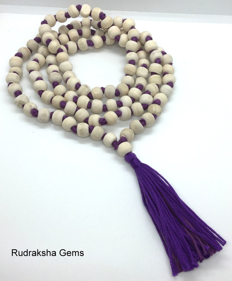 May include: A long necklace featuring natural light-coloured wooden beads strung on a vibrant purple cord, arranged in multiple loops. The necklace terminates in a long, matching purple thread tassel. The text Rudraksha Gems appears at the bottom.