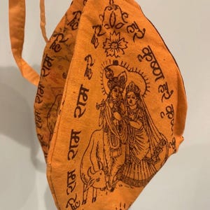 Puede incluir: Mascarilla facial de tela naranja con una ilustración marrón de deidades hindúes y escritura. La mascarilla tiene una correa y está hecha de un material suave y texturizado. La ilustración representa una pareja con una vaca.