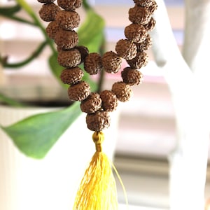 Peut inclure: Un collier de perles mala brun avec un pompon jaune. Les perles sont rondes et ont une texture rugueuse. Le pompon est fait de fines mèches de fil.