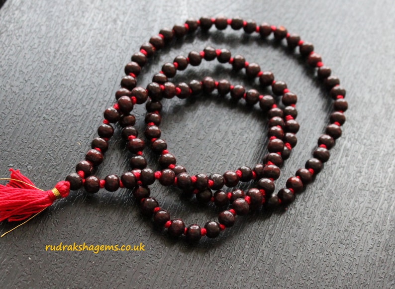 May include: A dark brown wooden mala with red string and a red tassel. The mala has 108 beads and is used for meditation and prayer.