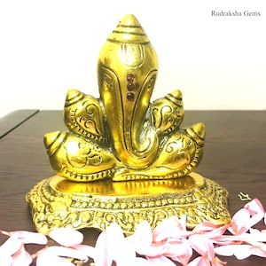 Puede incluir: Una estatua dorada de Ganesh con detalles intrincados. La estatua presenta una cabeza grande, orejas prominentes y un tronco. Tres gemas rojas adornan la frente. La estatua se asienta sobre una base ornamentada. El texto "Rudraksha Gems" est&aacute; en la esquina superior derecha.