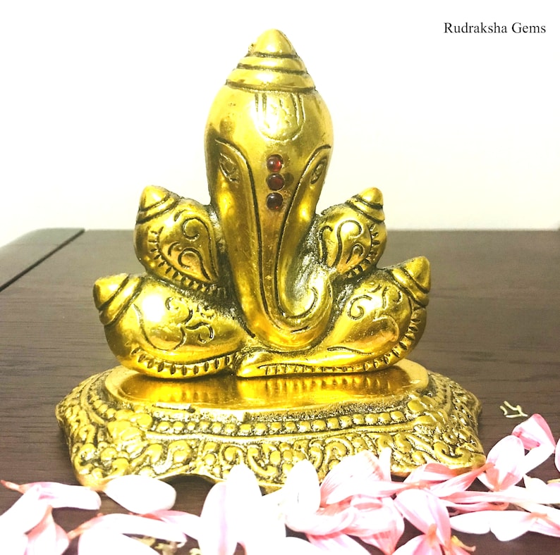 Puede incluir: Una estatua dorada de Ganesh con detalles intrincados. La estatua presenta una cabeza grande, orejas prominentes y un tronco. Tres gemas rojas adornan la frente. La estatua se asienta sobre una base ornamentada. El texto "Rudraksha Gems" est&aacute; en la esquina superior derecha.