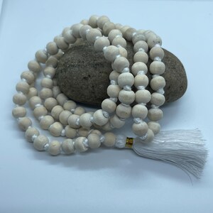 May include: A strand of light-coloured, matte wooden beads strung on white cord, arranged in a circular pattern. The beads are uniform in size and shape, featuring a small gold-coloured metal accent near a white tassel at the end of the strand.