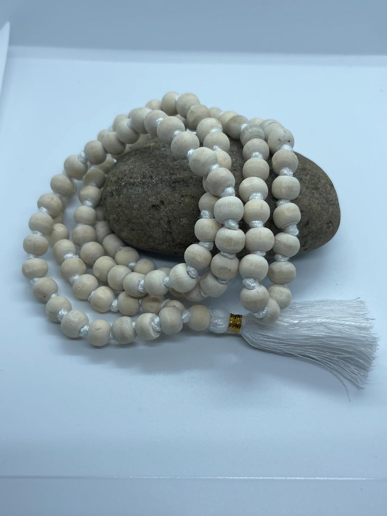 May include: A strand of light-coloured, matte wooden beads strung on white cord, arranged in a circular pattern. The beads are uniform in size and shape, featuring a small gold-coloured metal accent near a white tassel at the end of the strand.