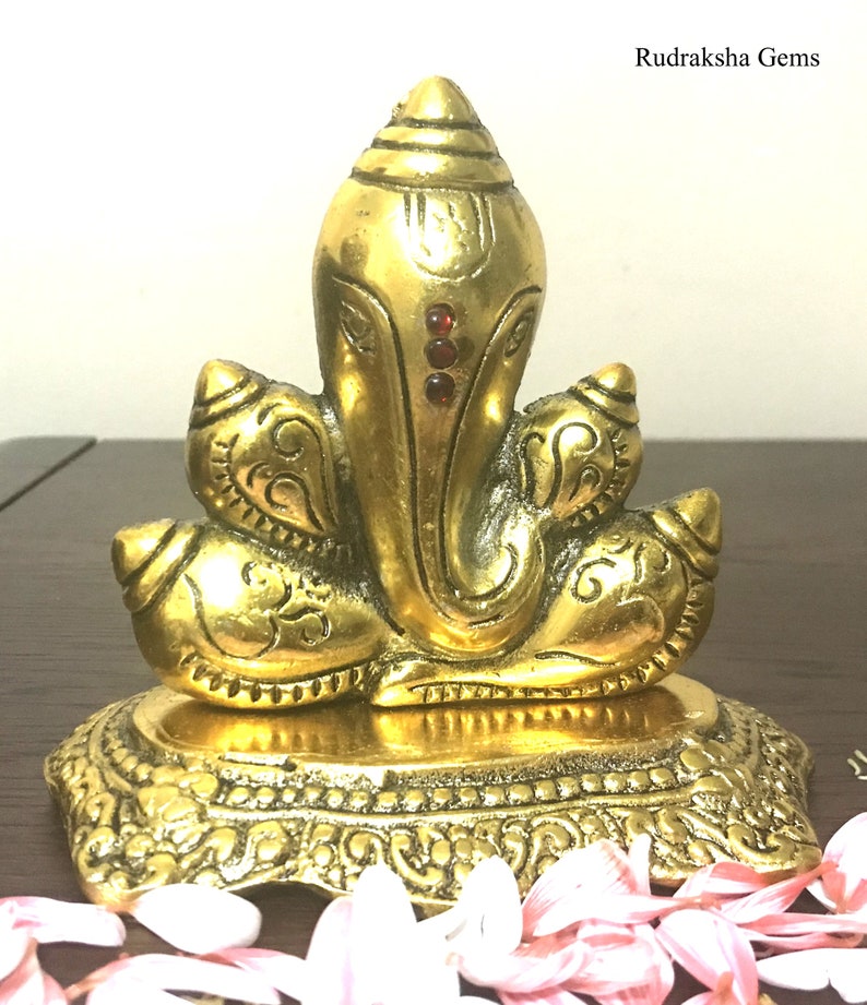 Puede incluir: Una estatua dorada de Ganesh de lat&oacute;n con detalles intrincados. La estatua presenta a Ganesh sentado con una cabeza grande, adornada con tres gemas rojas. La base es ornamentada y el dise&ntilde;o es muy detallado.