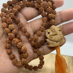 Peut inclure: Un mala en rudraksha marron avec un pendentif en bois sculpté et un pompon orange. Le mala est composé de 108 perles et est utilisé pour la méditation et la prière.