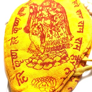 Puede incluir: Bolsa de tela amarilla con una imagen roja de una deidad y texto sánscrito. La bolsa tiene un cierre de cordón.