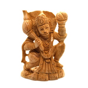 Puede incluir: Una talla de madera de la deidad hindú Hanuman, representada en posición de rodillas sosteniendo un garrote y una fruta. La talla es detallada e intrincada, con un color marrón cálido.