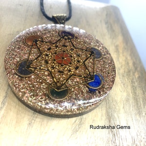 May include: A round pendant with a gold-colored Metatron's Cube design, set with colorful gemstones. The pendant is on a black cord. The background is a light-colored wooden surface. The text "Rudraksha Gems" is visible.