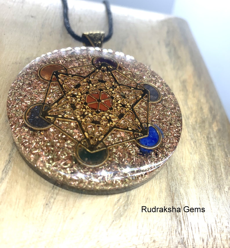 May include: A round pendant with a gold-colored Metatron's Cube design, set with colorful gemstones. The pendant is on a black cord. The background is a light-colored wooden surface. The text "Rudraksha Gems" is visible.
