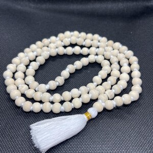 May include: A strand of light beige wooden beads strung on a white cord, arranged in a circular pattern. The necklace features a white fabric tassel attached with a small gold-coloured metallic band at the base of the bead loop.