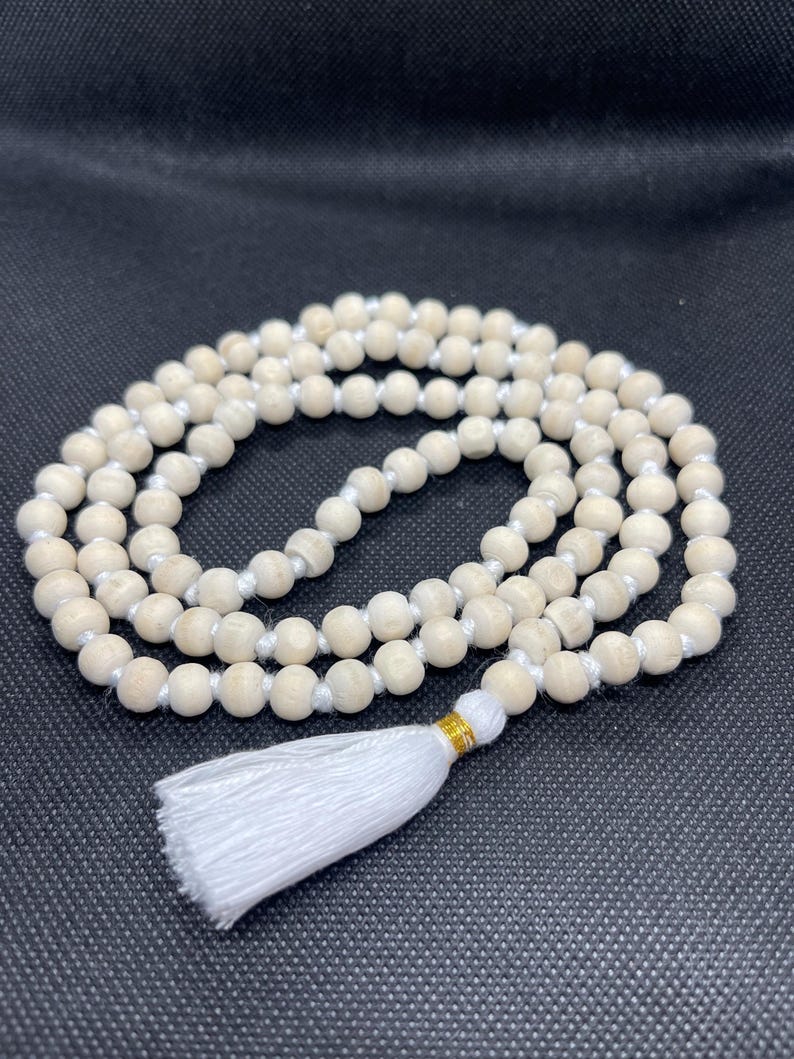 May include: A strand of light beige wooden beads strung on a white cord, arranged in a circular pattern. The necklace features a white fabric tassel attached with a small gold-coloured metallic band at the base of the bead loop.