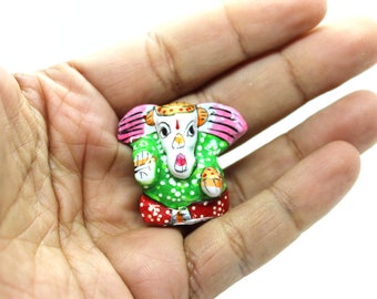 May include: A small, hand-painted figurine of the Hindu deity Ganesha. The figurine is green with white and red accents, and has a pink head with a white face. The figurine is sitting on a hand.