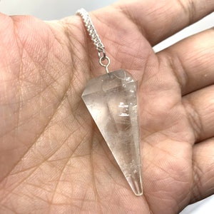 May include: A clear quartz crystal pendulum with a silver chain. The pendulum is shaped like a point and is hanging from a silver chain.