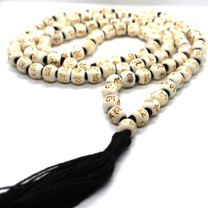 K&ouml;nnte beinhalten: Wei&szlig;e Holz-Mala-Perlen mit einem schwarzen Quasten. Die Perlen sind mit dem Sanskrit-Wort "Ram" in goldener Schrift versehen.