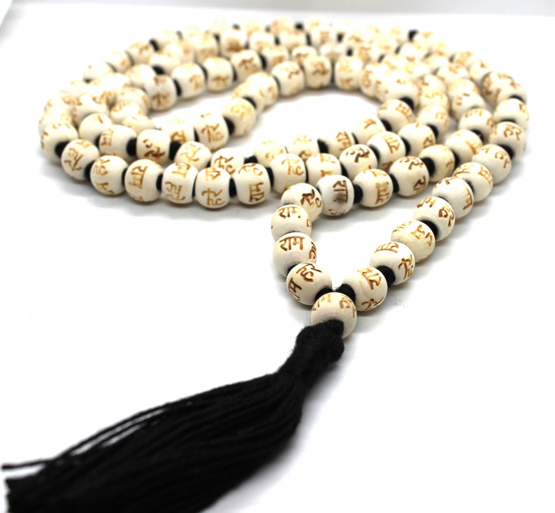 K&ouml;nnte beinhalten: Wei&szlig;e Holz-Mala-Perlen mit einem schwarzen Quasten. Die Perlen sind mit dem Sanskrit-Wort "Ram" in goldener Schrift versehen.