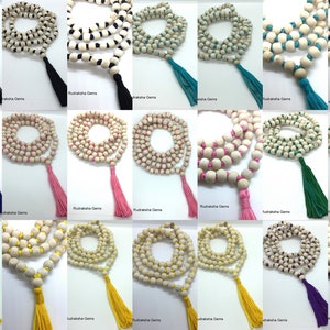 May include: A collection of twenty-four mala bead necklaces arranged in a grid. Each necklace consists of light-coloured wooden beads strung together with coloured thread, featuring a matching fabric tassel at the end of each strand.