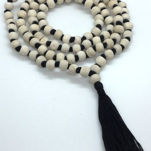 May include: A long strand of round, light-coloured wooden beads strung on a black cord, arranged in multiple loops. The necklace features a prominent black tassel at the end and includes the text Rudraksha Gems printed in the bottom left corner.