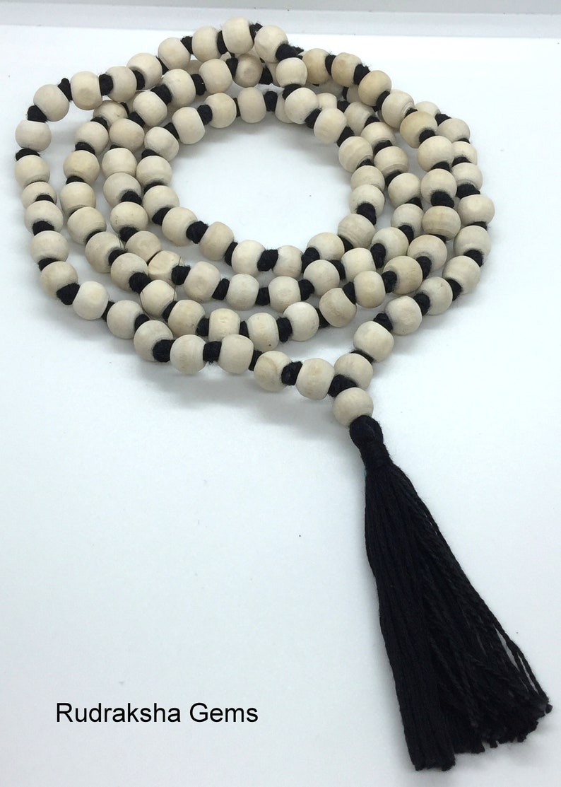 May include: A long strand of round, light-coloured wooden beads strung on a black cord, arranged in multiple loops. The necklace features a prominent black tassel at the end and includes the text Rudraksha Gems printed in the bottom left corner.