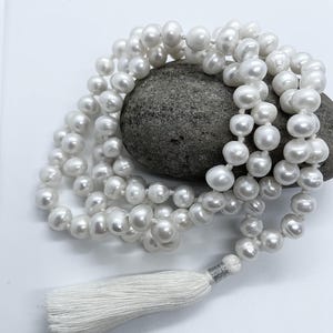 Puede incluir: Un collar de perlas blancas con una borla. El collar está drapeado sobre una piedra gris. Las perlas son redondas y tienen un brillo lustroso. La borla está hecha de hilo blanco y está unida al collar con metal plateado.