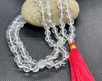 Puede incluir: Un mala de cuarzo transparente con un borla roja. El mala está hecho de cuentas redondas y está enhebrado en un cordón blanco. La borla está hecha de hilo rojo y tiene un acento dorado.