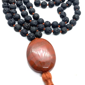May include: A black lava stone mala necklace with a red jasper pendant and a tassel. The necklace is made of 108 beads and is approximately 18 inches long.