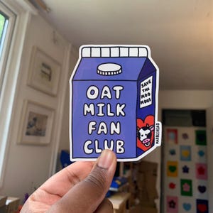 Pode incluir: Um autocolante azul em forma de embalagem de leite de aveia com as palavras "OAT MILK FAN CLUB" em branco. Uma ilustração de vaca está na lateral com o texto "SAVE THE MOOS". O autocolante é segurado por uma mão.