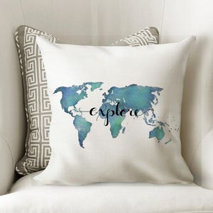 K&ouml;nnte beinhalten: Ein wei&szlig;es Kissen mit einem Aquarell-Weltkarten-Design in Blaut&ouml;nen und Gr&uuml;n. Das Wort "explore" ist in schwarzer Kursivschrift &uuml;ber die Karte geschrieben.