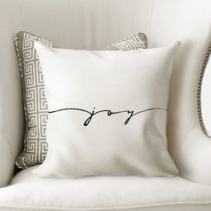 May include: A white decorative pillow with the word "joy" in a black script. The pillow is on a white armchair with a patterned pillow behind it. The chair has a tall back and is in a well-lit room.