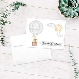 Puede incluir: Un globo aerost&aacute;tico blanco con una jirafa y un le&oacute;n dentro, volando sobre un sol amarillo con una cara sonriente. La pancarta dice "Welcome Liam James".