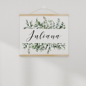 May include: A white canvas print with a wooden frame, featuring a green watercolor floral design and the name "Juliana" in black calligraphy.