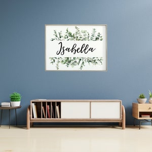 May include: A framed print with a white background and a green watercolor illustration of eucalyptus leaves. The name "Isabella" is written in black cursive script on a white banner.