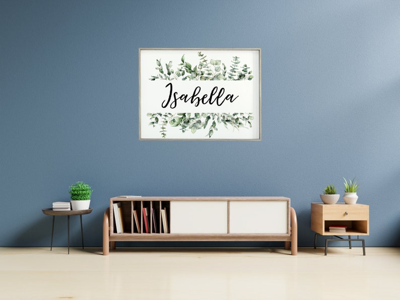 May include: A framed print with a white background and a green watercolor illustration of eucalyptus leaves. The name "Isabella" is written in black cursive script on a white banner.