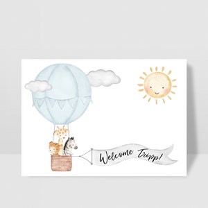 Puede incluir: Una ilustraci&oacute;n de acuarela de un globo aerost&aacute;tico con una jirafa, una cebra y un le&oacute;n dentro. El globo es azul y blanco con una pancarta que dice "&iexcl;Bienvenido Tripp!" Hay un sol amarillo con una cara sonriente en la esquina superior derecha.