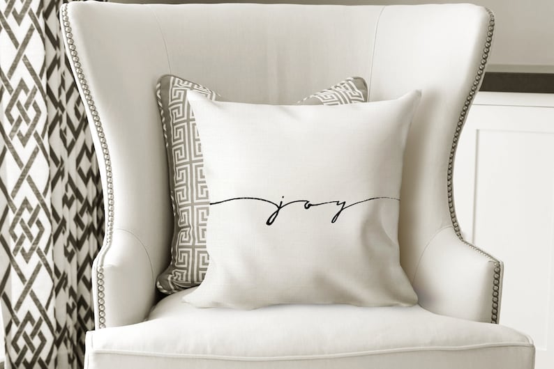 May include: A white decorative pillow with the word "joy" in a black script. The pillow is on a white armchair with a patterned pillow behind it. The chair has a tall back and is in a well-lit room.