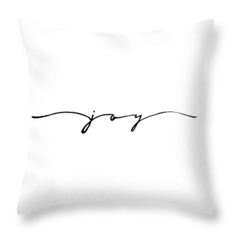 May include: White square throw pillow with the word "joy" written in a black, cursive font. The word is centered horizontally and sits on a single, thin, curved line.