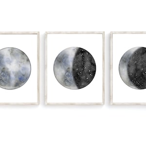 May include: Set of three framed watercolor paintings depicting different phases of the moon. Each painting features a circular moon with a gray and blue color scheme, with the middle painting showing a half moon with a black and white starry sky.