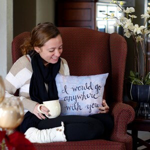 May include: A decorative pillow with the text "I would go anywhere with you" in black script. The pillow has a light blue background with a map design. The person is sitting in a red armchair.