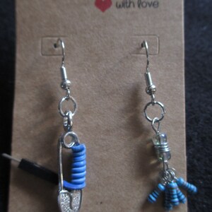 May include: A pair of handmade earrings. One earring features a silver safety pin charm with blue coiled wire. The other earring features a silver charm with a clear crystal bead and blue resistor beads.