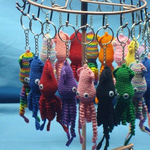 May include: A collection of crocheted squid keychains in various colors, including pink, blue, green, red, and purple. Each keychain has large, white eyes and is attached to a silver key ring.
