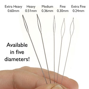 May include: Five different sizes of beading needles, held in a hand. The needles are made of metal and have a loop at the end. The sizes are labeled as Extra Heavy 0.60mm, Heavy 0.51mm, Medium 0.36mm, Fine 0.30mm, and Extra Fine 0.24mm. The text "Available in five diameters!" is also visible.