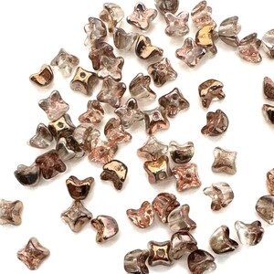 Op de afbeelding: Een verzameling kleine, transparante glazen kralen met een metallic gouden en rosé gouden afwerking. De kralen zijn vierpuntig stervormig met een gat in het midden.