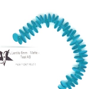 May include: A strand of teal blue glass lentil beads, 6mm in size, with a matte finish. The beads are arranged in a semi-circle shape. The text "Lentils 6mm Matte - Teal AB" is visible on a white label.